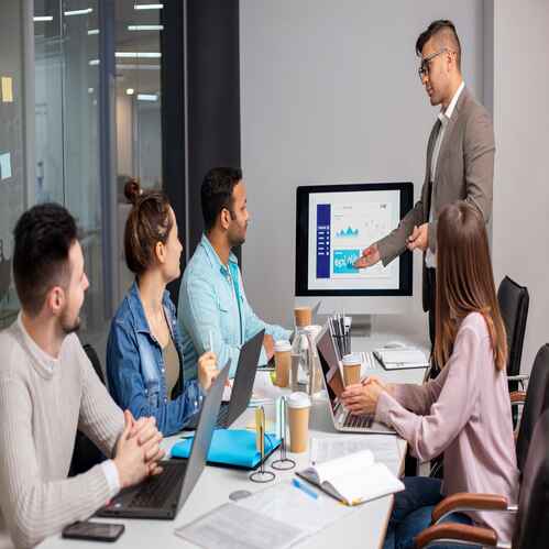 A Man In A Suit Presents Data On A Screen In A Conference Room To Four Attentive Colleagues