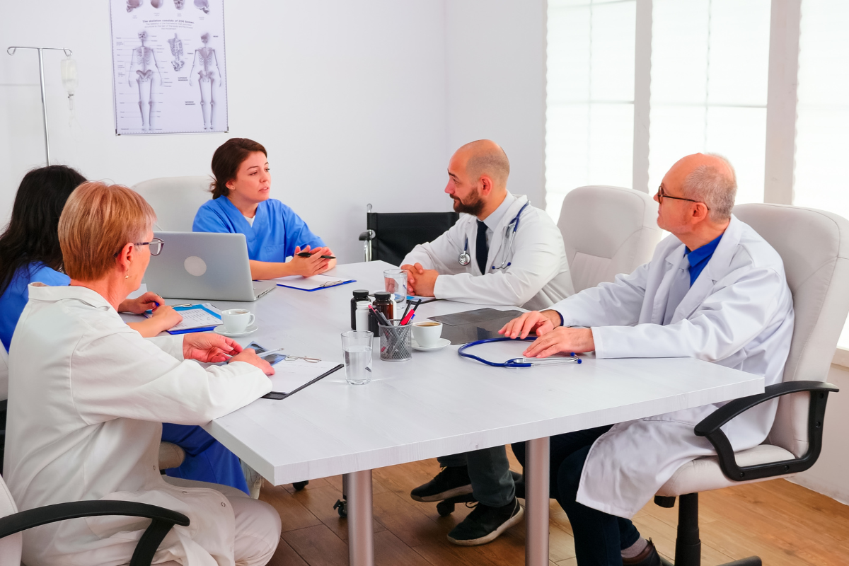 A Group Of Doctors Engaged In Discussion Around A Conference Table, Sharing Ideas And Collaborating On Patient Care