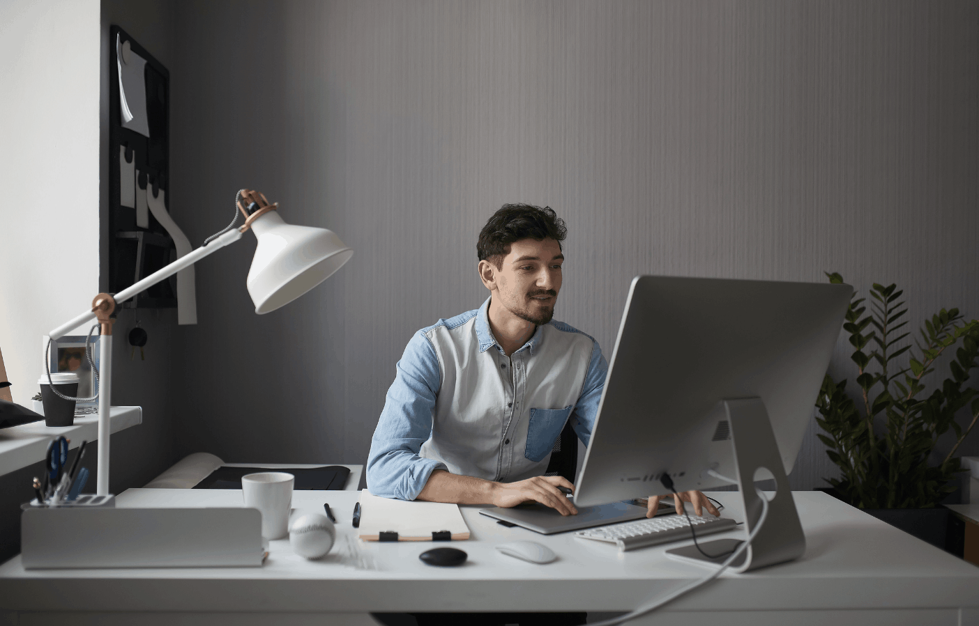 A Man Seated At A Desk, Working On A Computer With A Lamp Illuminating The Workspace